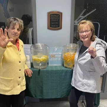 two women waving at the camera in front of drink dispensers filled with lemon and orange slices