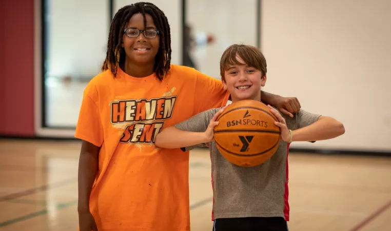 2 boys pose together smiling in the gymnasium, while the one on the right holds a basketball