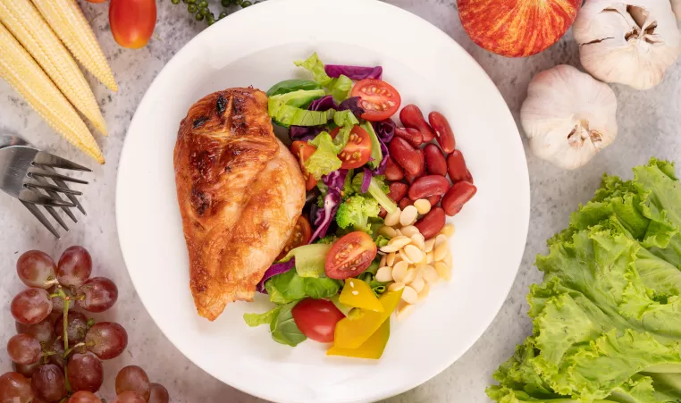 Plate full of healthy food items, including chicken, tomatoes, lettuce, peppers, and radish. It is surrounded by corn, forks, grapes, garlic, and lettuce on the surface holding the plate
